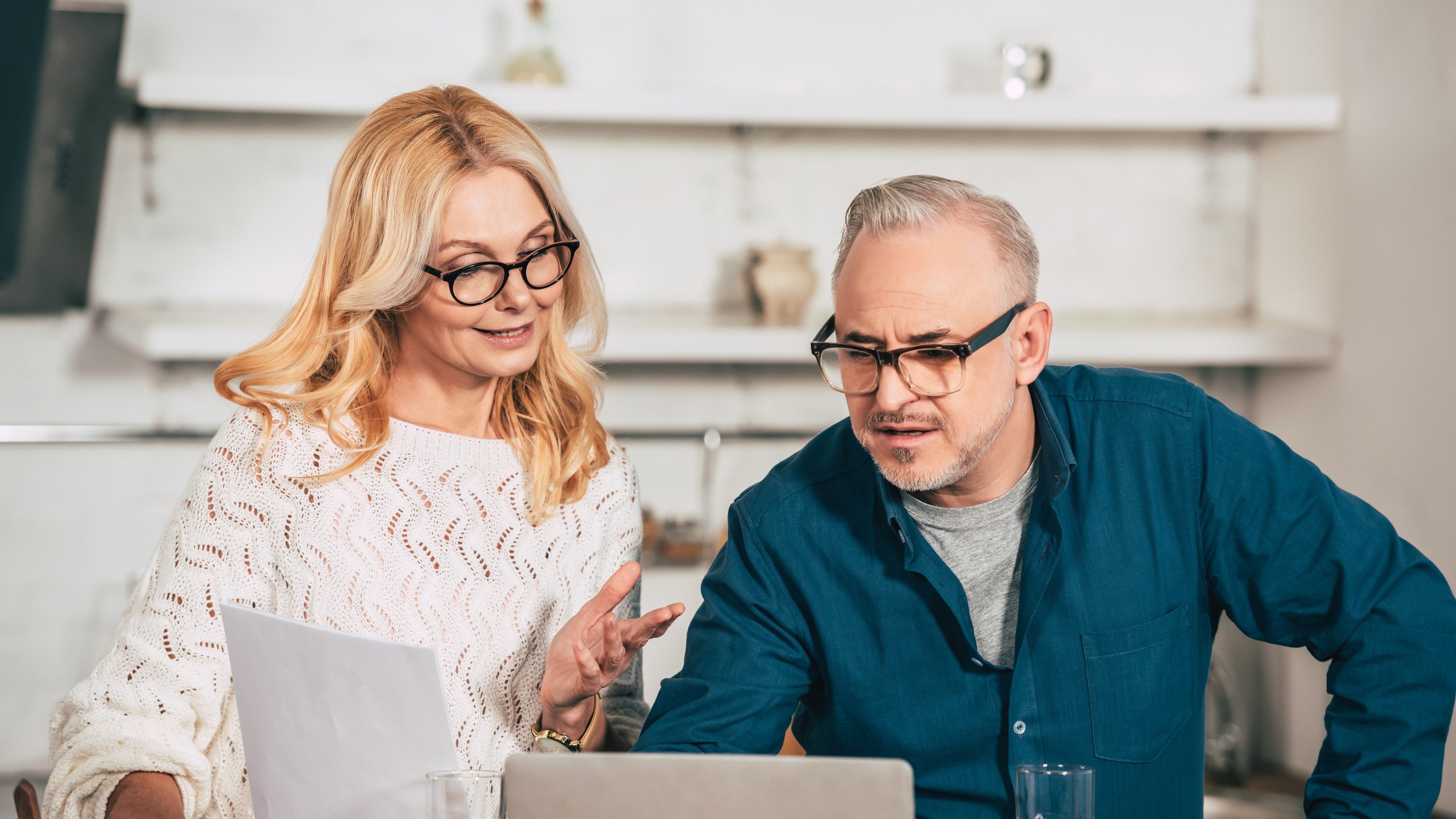 Eine blonde, langhaarige Frau mit Brille und ein grauhaariger Mann mit Brille sitzen vor einem geöffneten Laptop und diskutieren fröhlich.