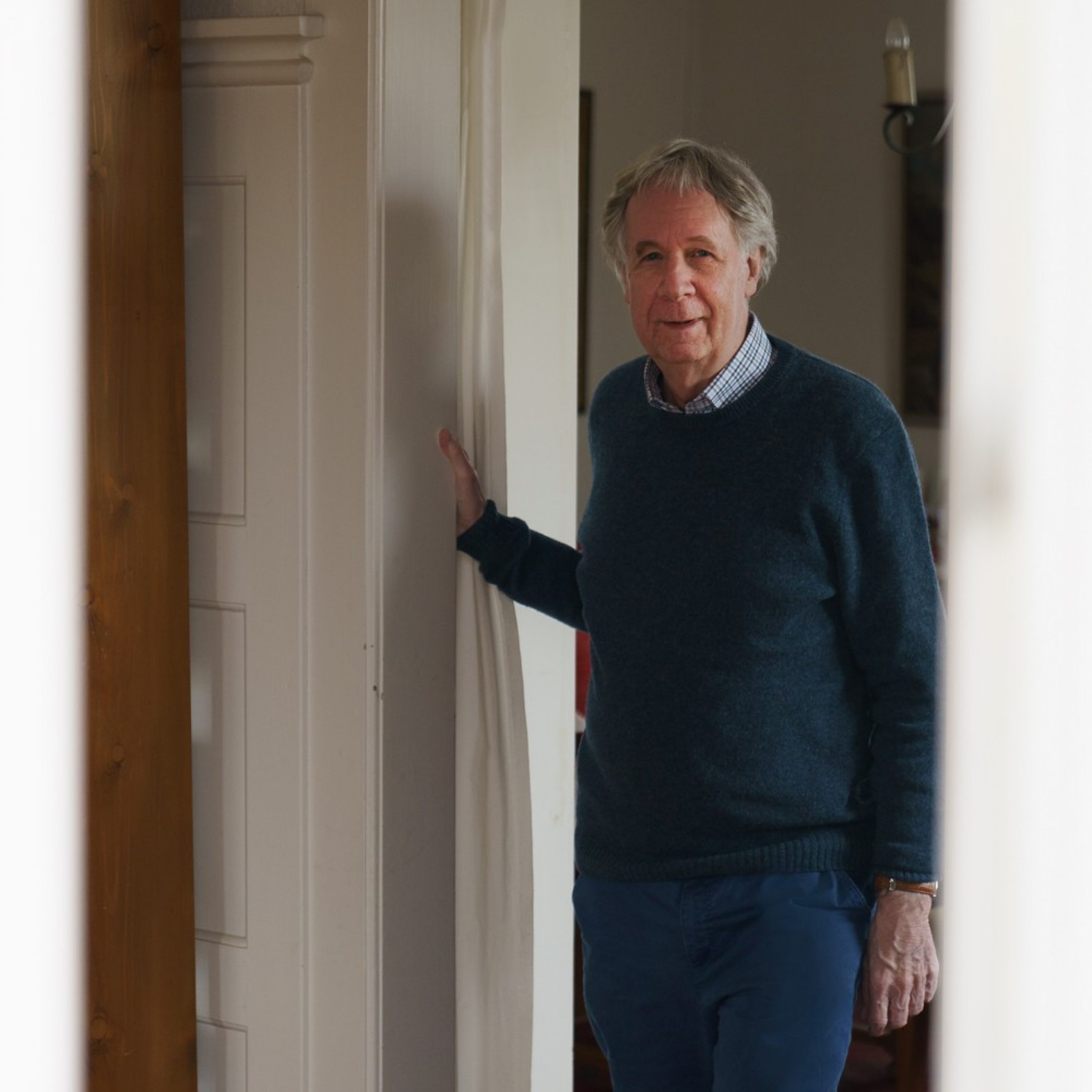 An older man stands by a door frame and looks into the camera. He’s wearing a shirt with a blue sweater over it.