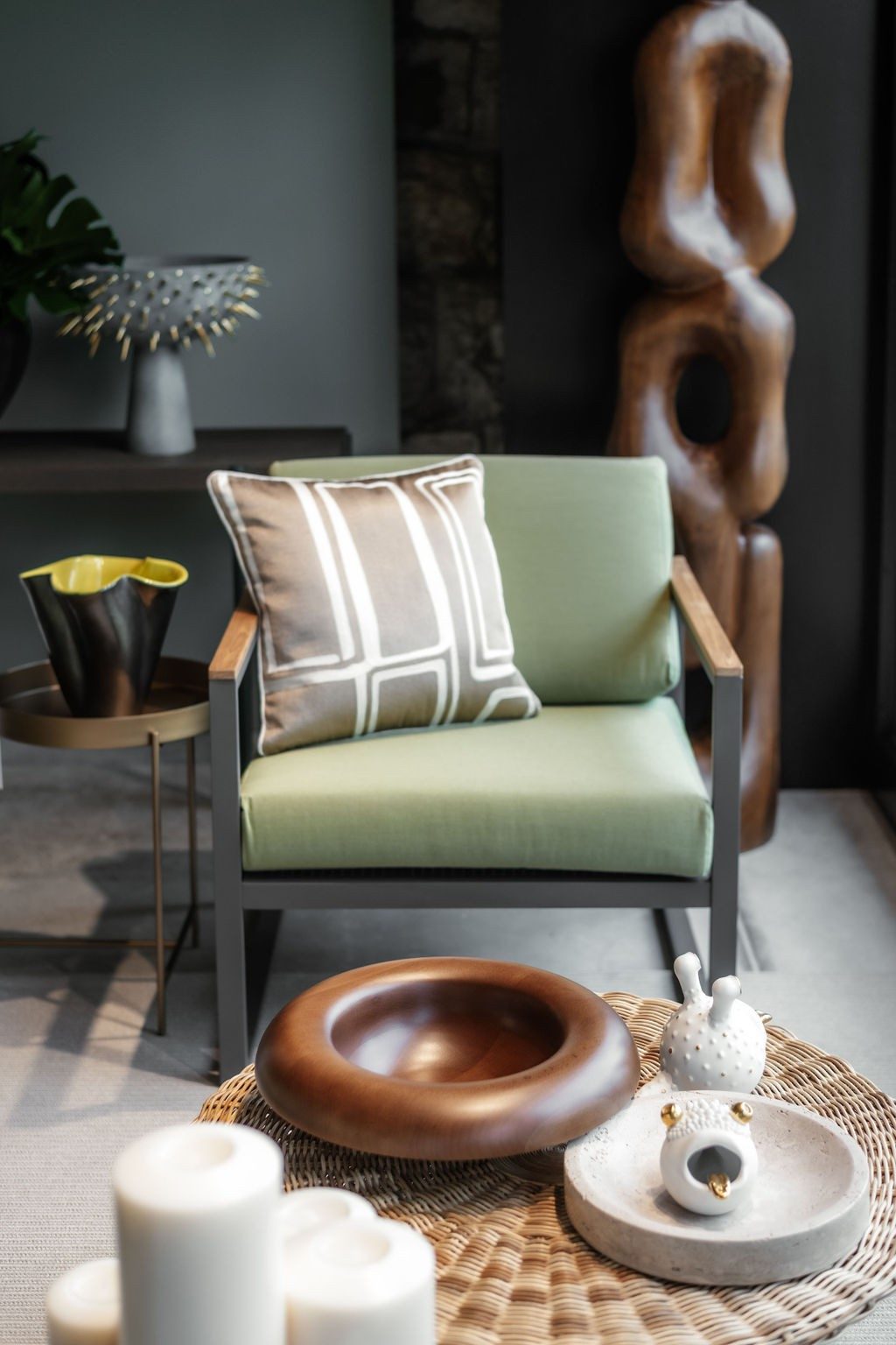 A green armchair with a cushion stands behind a round, braided coffee table. On the table are bowls and decorative items. One of the decorative items looks like a frog head with a golden tongue and golden eyes. Behind the armchair are two decorative items: a chain-like wooden sculpture and a decorative element with pointed elements.