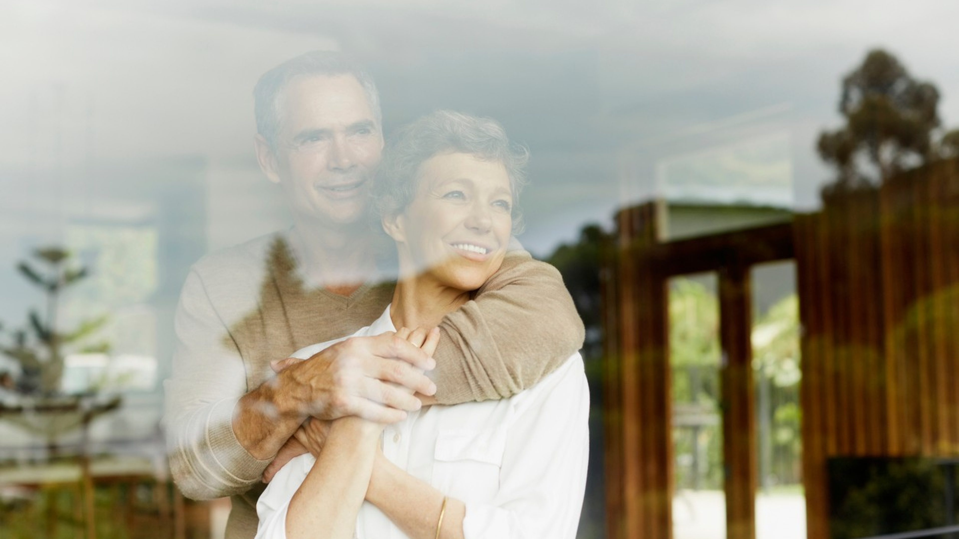 A couple in their mid-50s stands behind a windowpane hugging each other and looking out of the window. The colours are warm. The man is wearing a beige sweater, the woman a white blouse. Both are smiling and looking through the window into the distance.