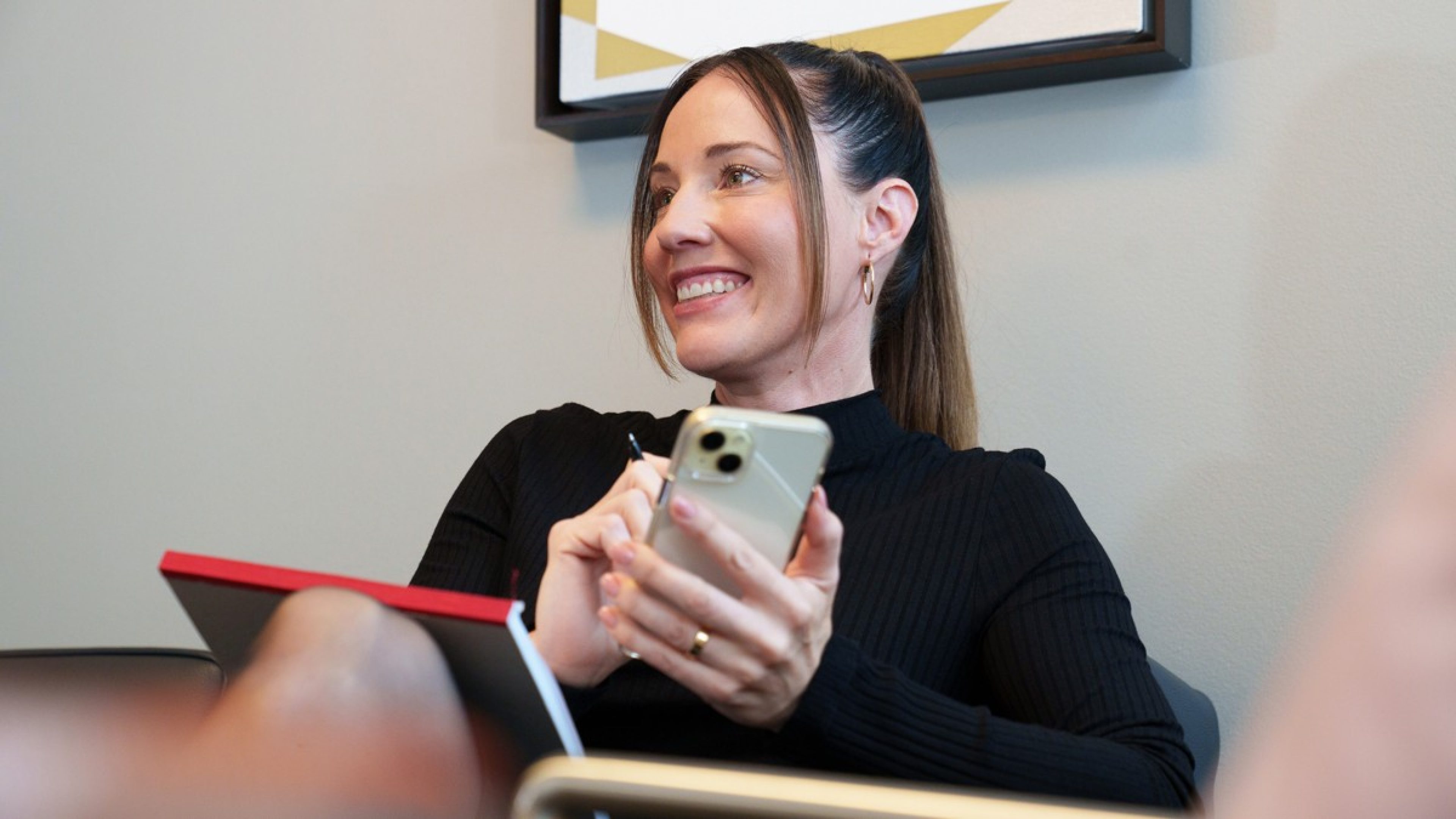 A woman with a ponytail looks to the side and laughs. She holds a mobile phone in her hand and has a notepad on her lap.