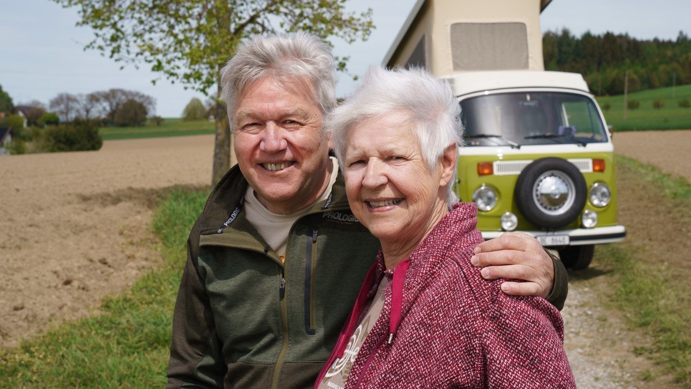 Kurt has his left arm around Monica while they look into the camera with a smile in front of their moss-green VW van. They are wearing dark green and dark red sweaters. 