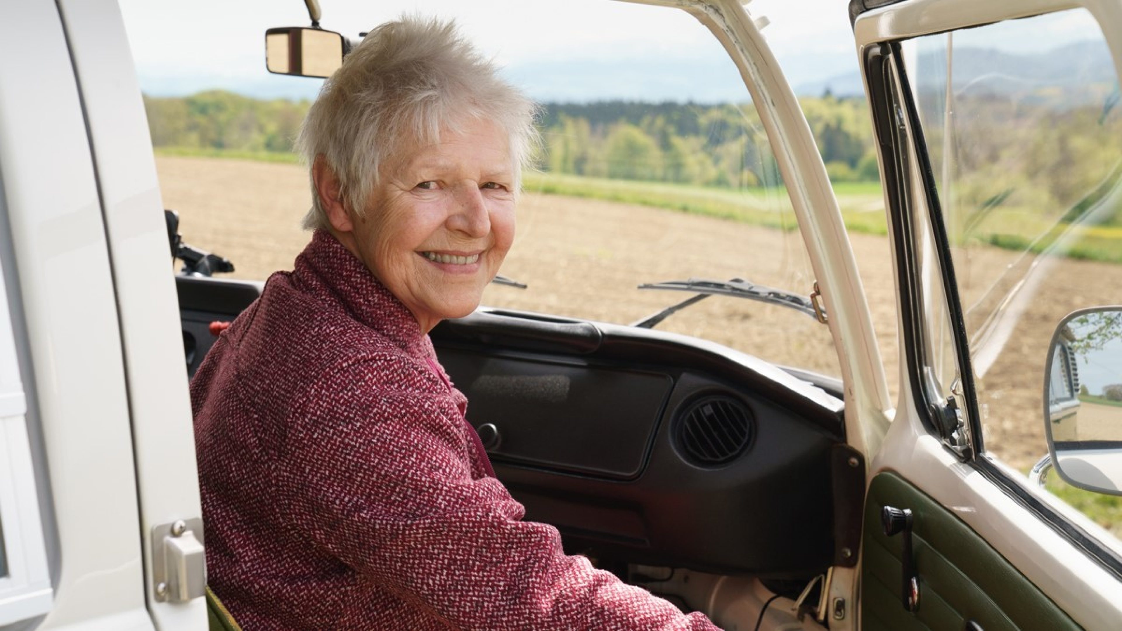 Monica sits in the passenger seat and smiles over her shoulder at the camera while her hand is on the door handle of the open car door. It seems as if she is going to embark on her next journey at any moment. She is wearing a burgundy sweater and in the background there is a field and a forest. 