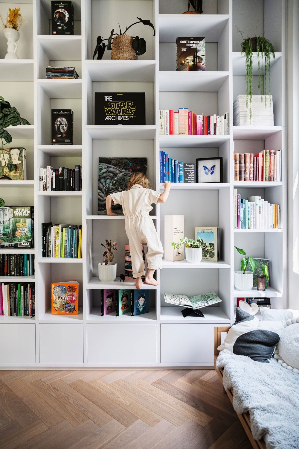 A child in a playsuit climbs up bookshelves. The library houses stylishly arranged books and Star Wars merchandise.