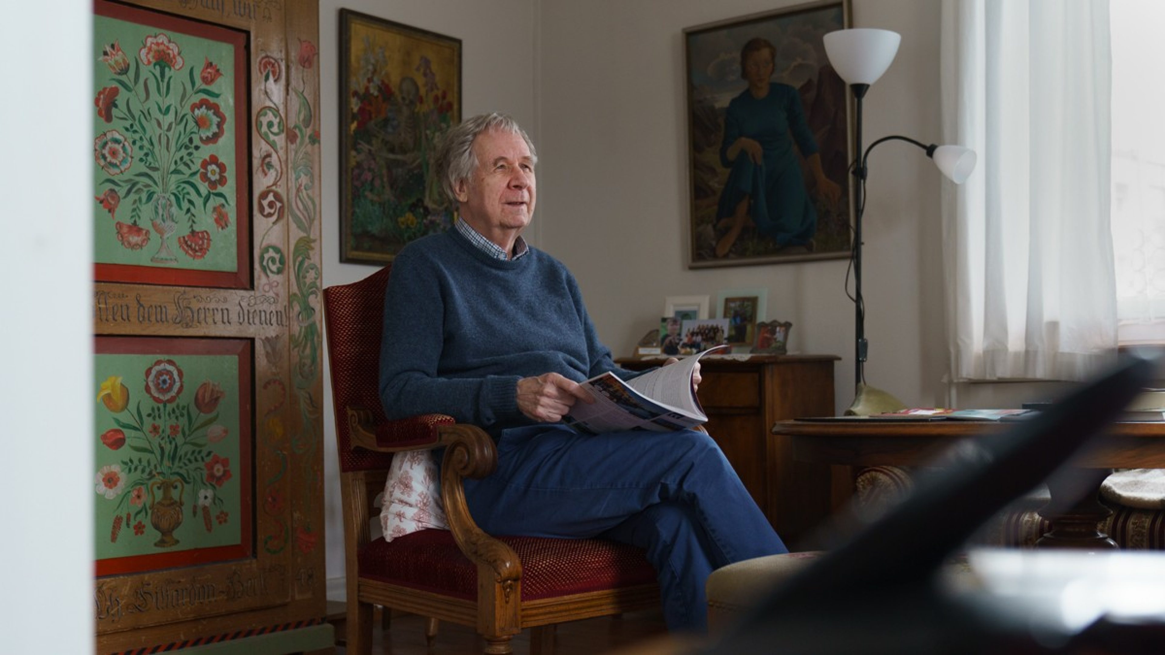 Ein älterer Mann sitzt in einem traditionell eingerichteten Wohnzimmer und liest eine Zeitschrift. Er trägt einen blauen Pullover und eine dunkelblaue Hose. 