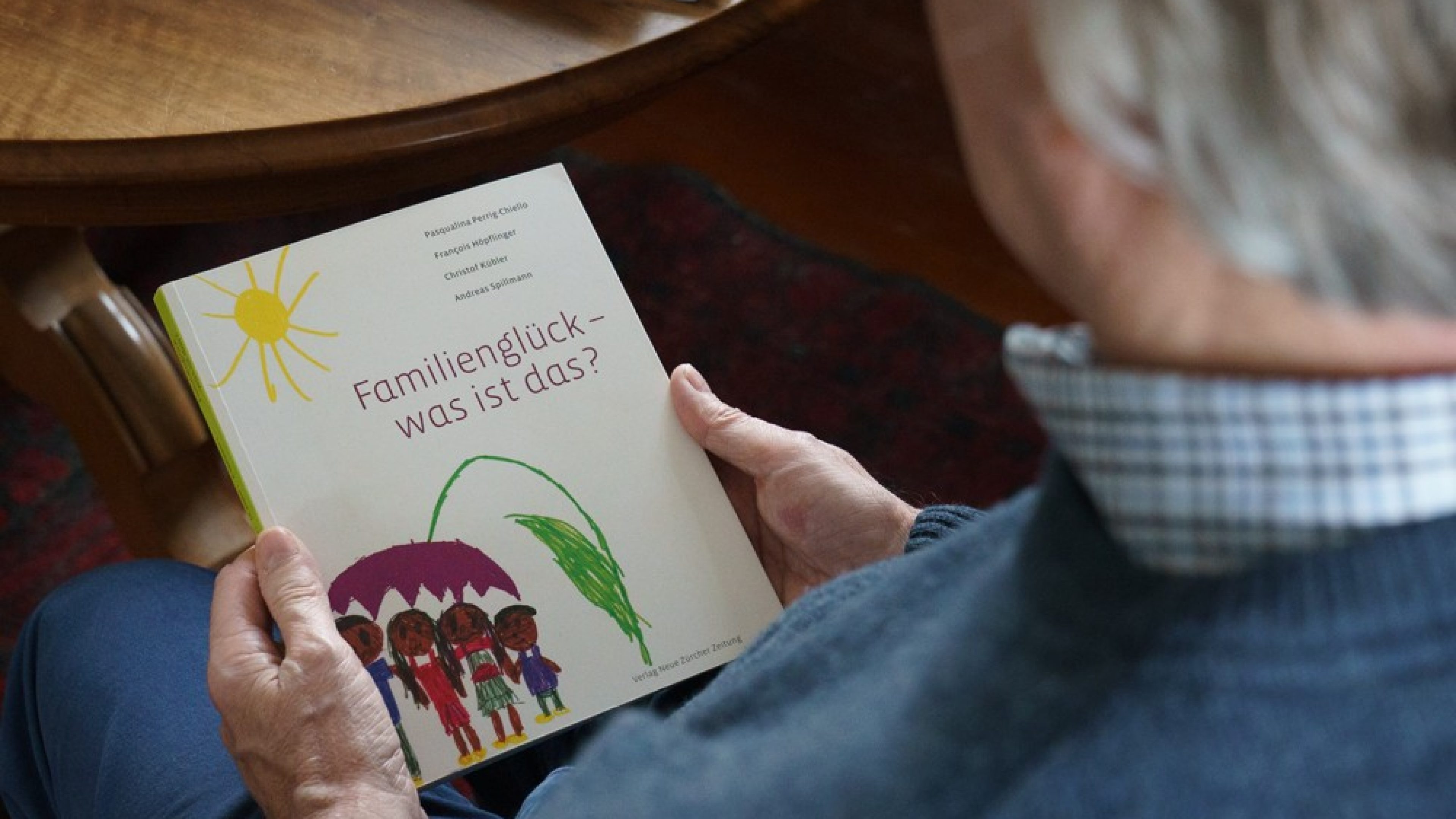 Vue par-dessus l’épaule d’un homme âgé. Celui-ci tient un livre intitulé: «Familienglück - was ist das» (Le bonheur familial – Qu’est-ce que c’est?). 