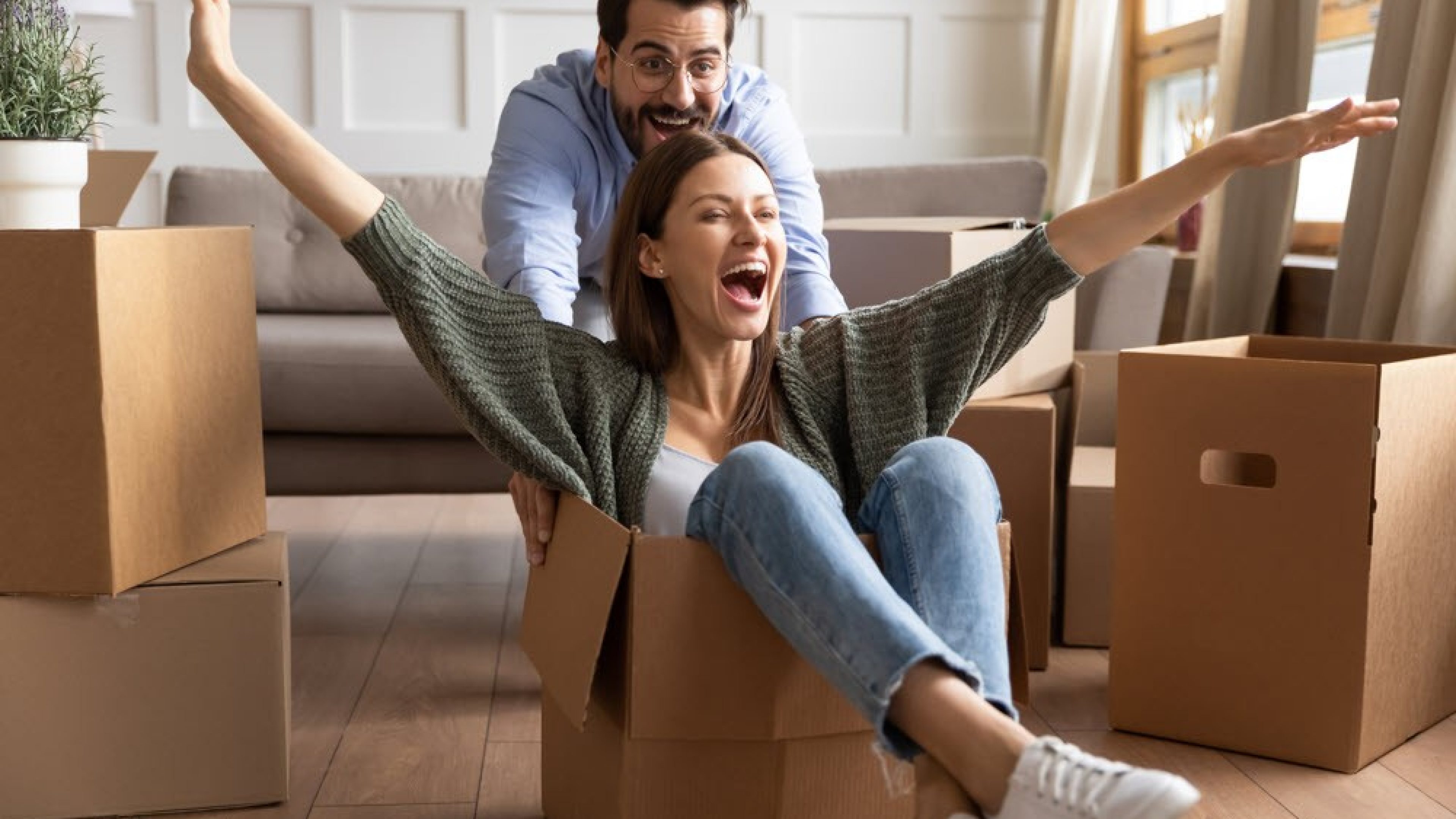 Une jeune femme souriante est assise dans un carton et son mari la pousse à travers le salon, dans lequel se trouvent de nombreuses cartons de déménagement.
