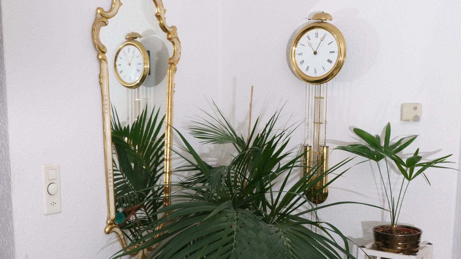 Ein Foto von Meinrad Grads Einrichtung zuhause mit einem altmodischen, vergoldeten Spiegel, einer Pendeluhr und Pflanzen in einer Ecke. Das Bild vermittelt ein Gefühl von Eleganz, Tradition und Naturverbundenheit in der Einrichtung.