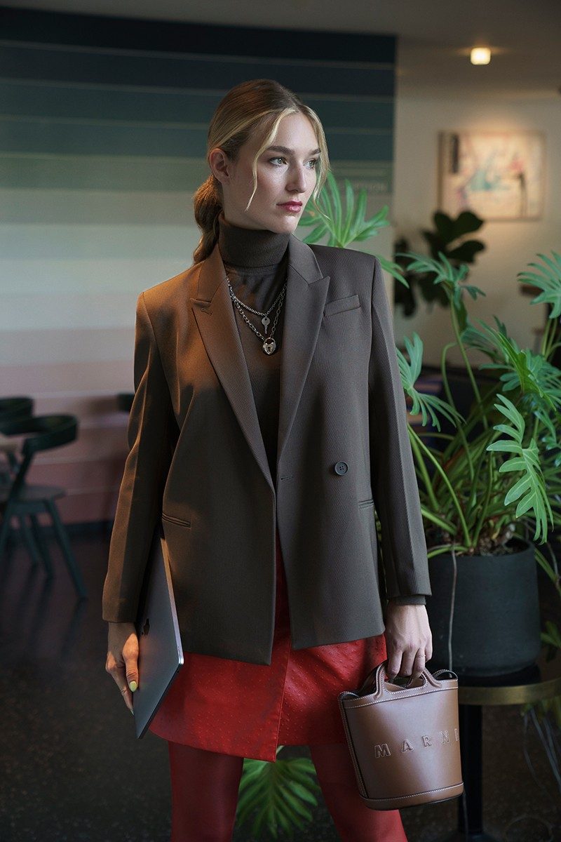 Manuela Frey, vêtue d’un blazer marron et d’un pull à col roulé, regarde par la fenêtre. 