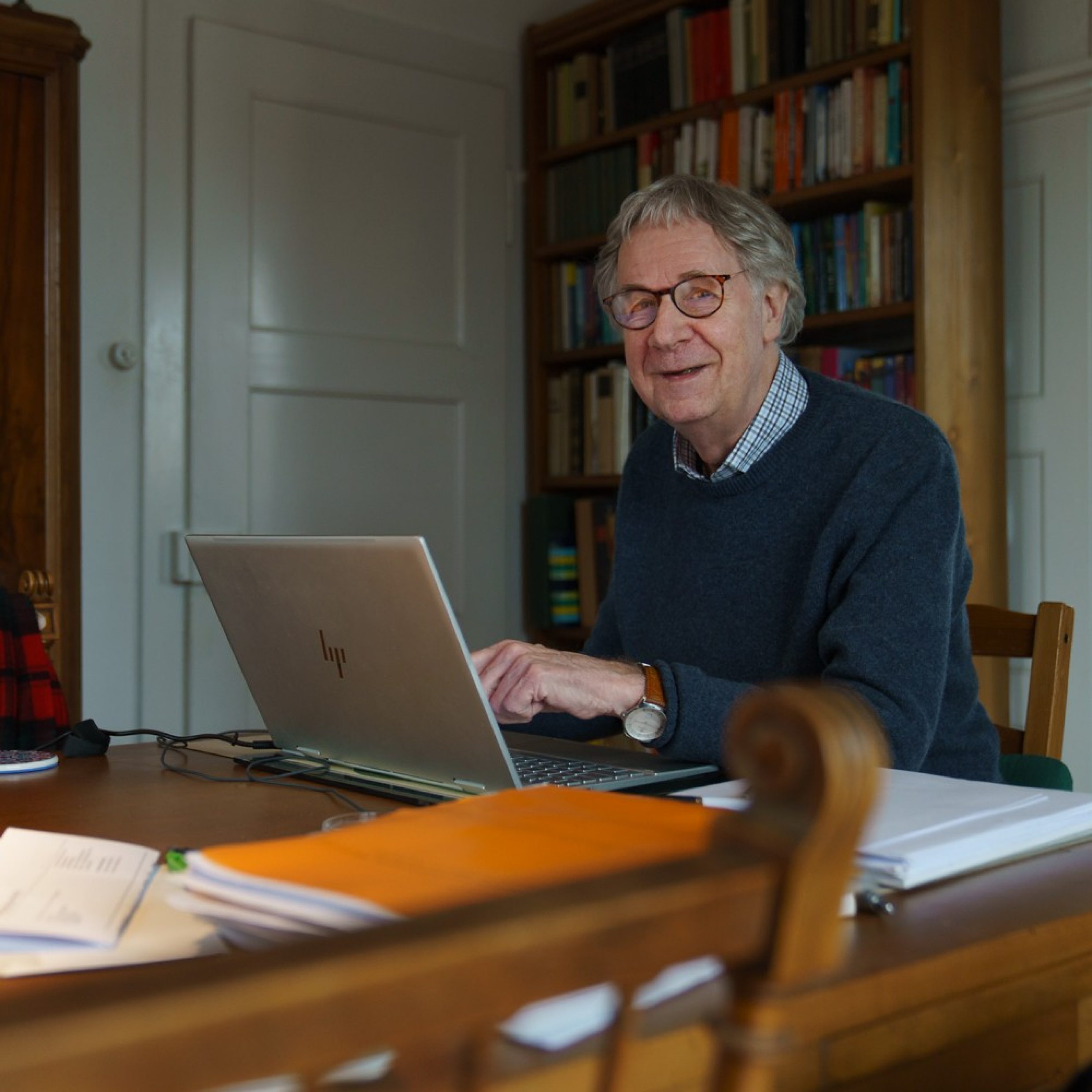 Un uomo anziano seduto a un tavolo davanti a un laptop mostra qualcosa sullo schermo. Sorride alla fotocamera. Ha i capelli grigi, porta gli occhiali e un maglione blu. 