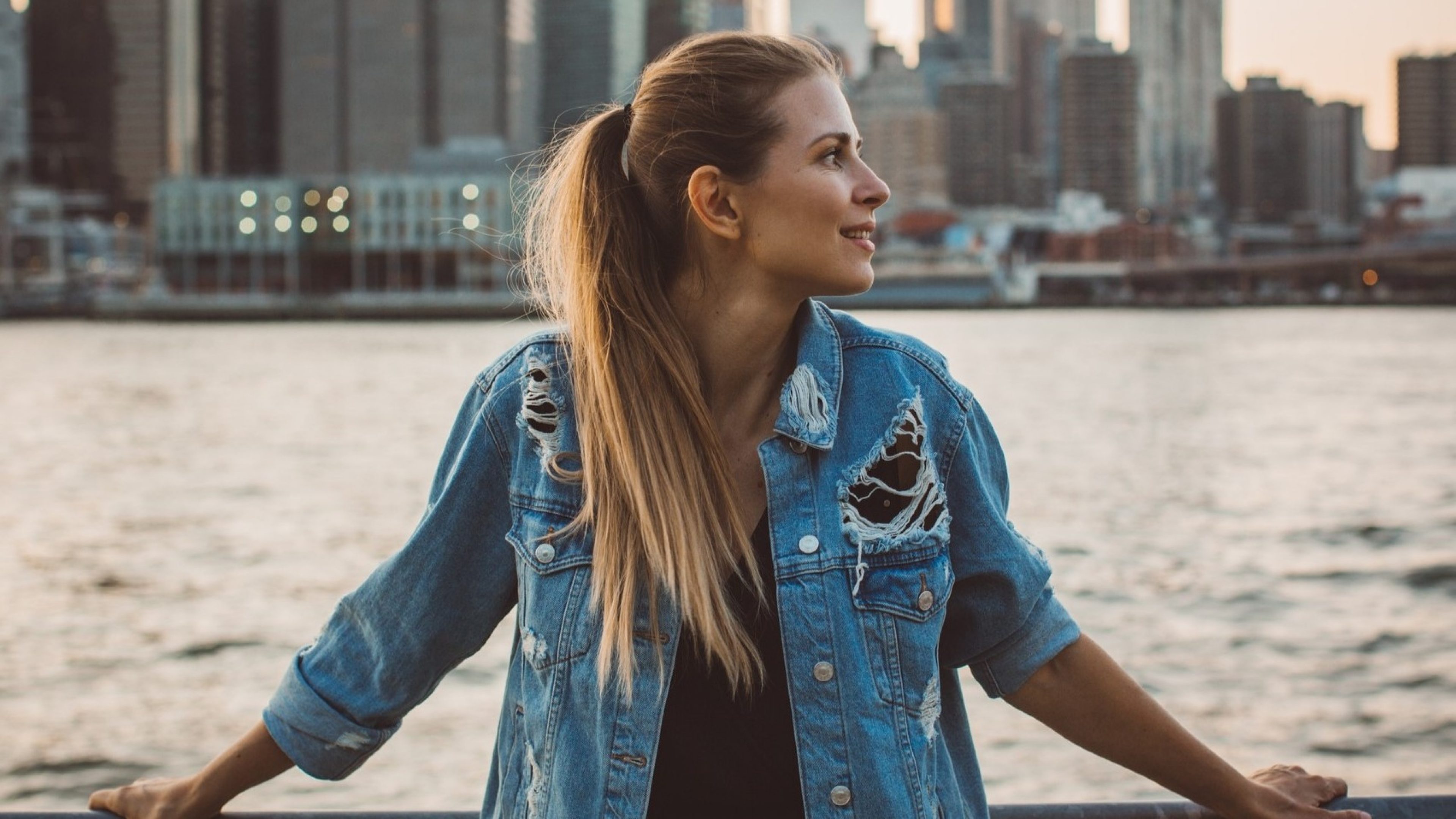 Una giovane donna dai lunghi capelli biondi e una giacca di jeans strappata si trova davanti a una ringhiera vicino all’acqua, con un paesaggio urbano sullo sfondo. Guarda di lato sorridendo, mentre il sole tramonta.